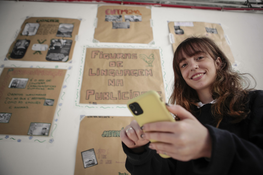 A foto foi tirada de baixo para cima. Na imagem está uma jovem, branca, de cabelos loiros, liso e comprido. Ela veste uma blusa preta. Está sorrindo, enquanto segura um celular amarelo em uma das mãos. Ao fundo, ha uma parede branca onde estão alguns cartazes colados com algumas frases escritas.
