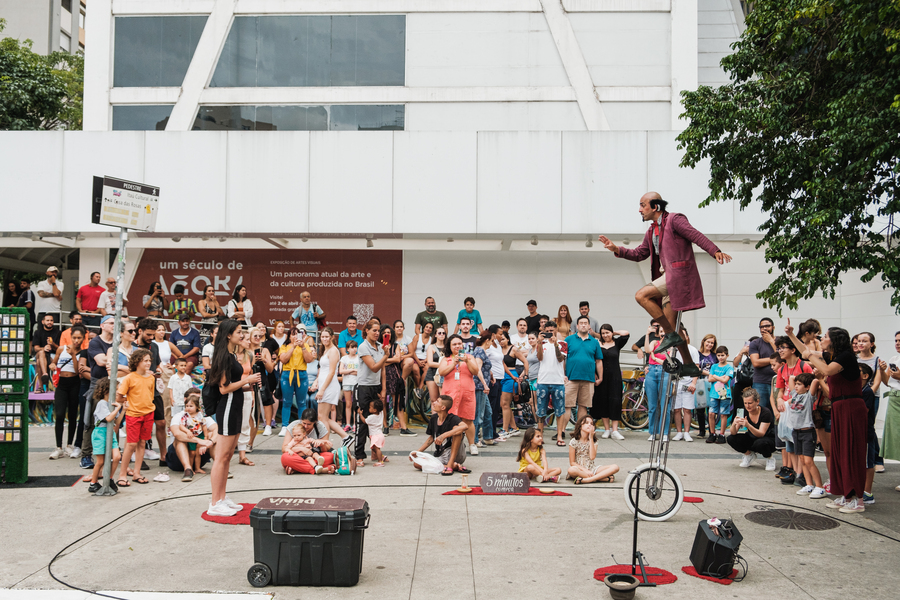 Na foto estão pessoas diversas reunidas em um espaço ao ar livre. Elas assistem a uma apresentação de um artista que está que equilibrando em um monociclo circense.